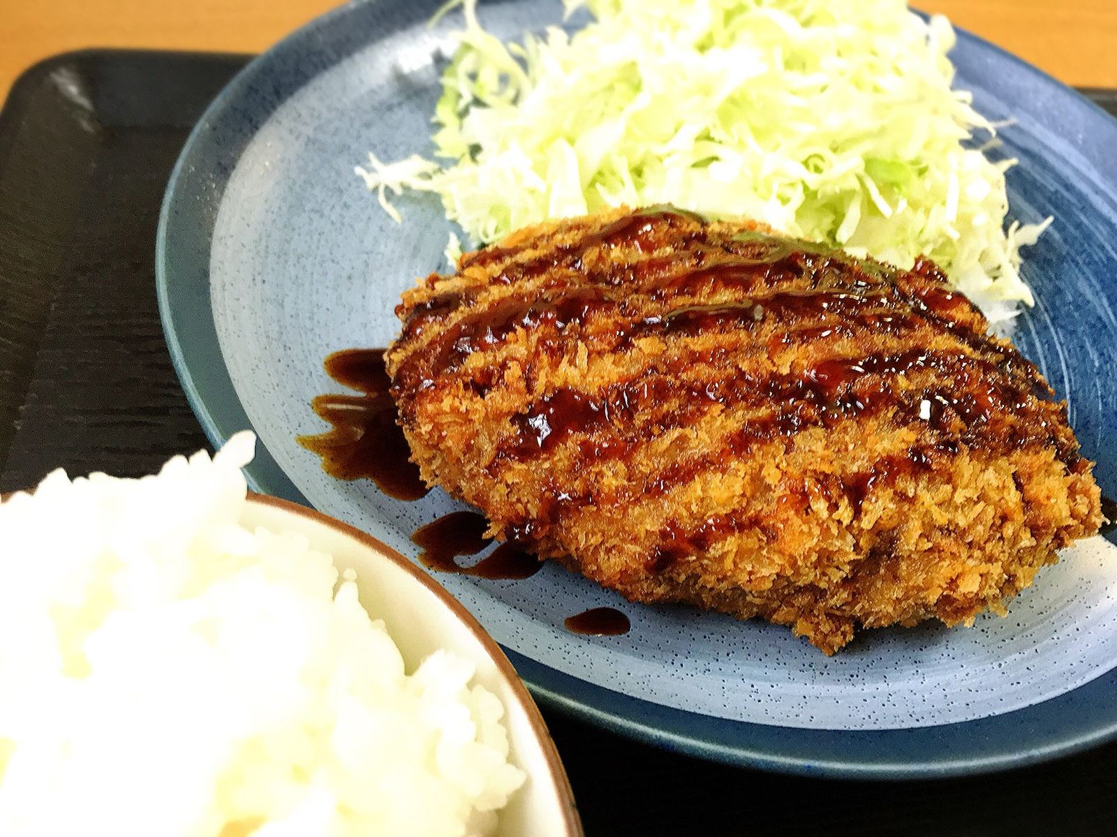 青い皿のメンチカツにソースをかけたところ。千切りキャベツとご飯