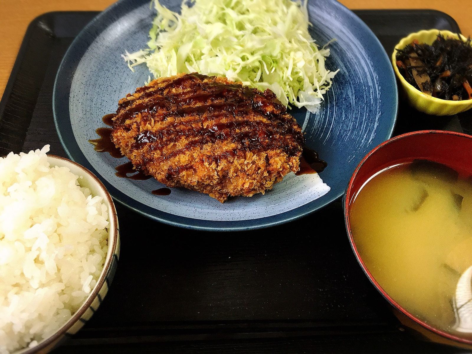 メンチカツ定食の全景。青い皿のメンチカツ、ご飯、味噌汁、小鉢をお盆にのせて